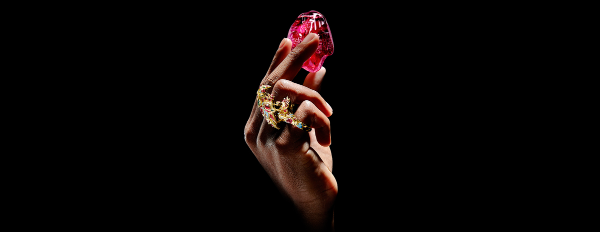 One hand emerges from the bottom of the photo wearing a golden bird-shaped ring covered in blue, red, and green jewels. The hand is delicately hoisting up and ruby stone with subtle carvings throughout it. 
